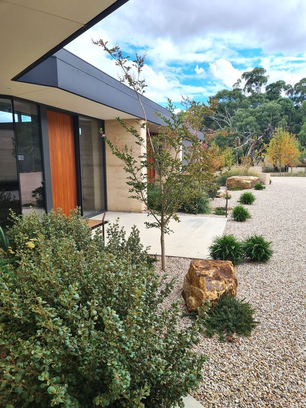 A contemporary single-storey residence viewed from a landscaped front courtyard. The home features a broad projecting roof canopy, dark-framed glazing, a tall warm timber entry door, pale masonry walls and a minimalist gravel garden with sculptural rocks, native-style planting and a concrete path. Trees and a bright sky are visible beyond the property.