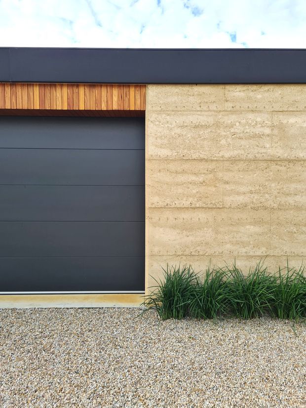 Close-up exterior detail of a contemporary facade featuring a large dark charcoal sectional garage door, warm vertical timber cladding above it, and a light textured masonry or concrete-block wall. Gravel landscaping and ornamental grasses line the base.
