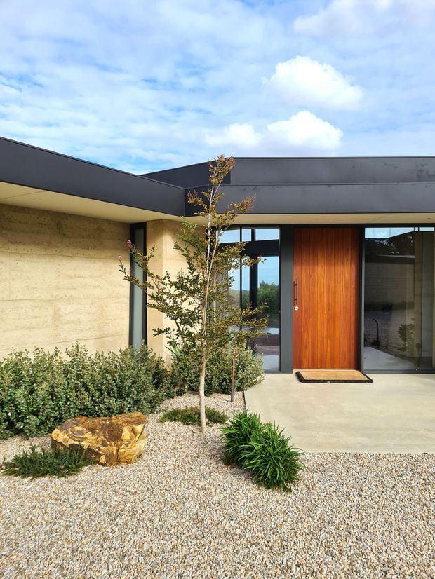 Front exterior view of a low-slung contemporary residence with a flat roof, dark metal fascia, warm textured masonry walls, extensive dark-framed glazing, and a tall timber entry door. A gravel landscape with shrubs, a young tree, ornamental grasses, and a feature rock leads to the covered entry.
