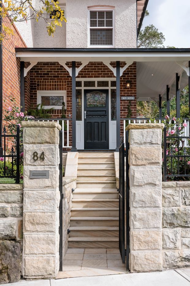 Front exterior view of a traditional two-storey brick residence, featuring a covered porch, decorative timber brackets, black-and-white detailing, stained-glass entry door, sandstone boundary pillars and steps, wrought-iron gates, and established planting.