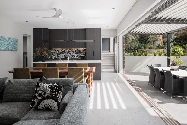 Open-plan contemporary living, dining and kitchen interior with a large grey sectional sofa in the foreground, a timber dining table, dark full-height kitchen cabinetry, and a stone island or counter. The room opens broadly to a furnished outdoor dining terrace through large retractable glazing, with strong linear sunlight and an integrated louvred overhead shade. Pale walls and flooring contrast with dark joinery and warm timber furnishings.