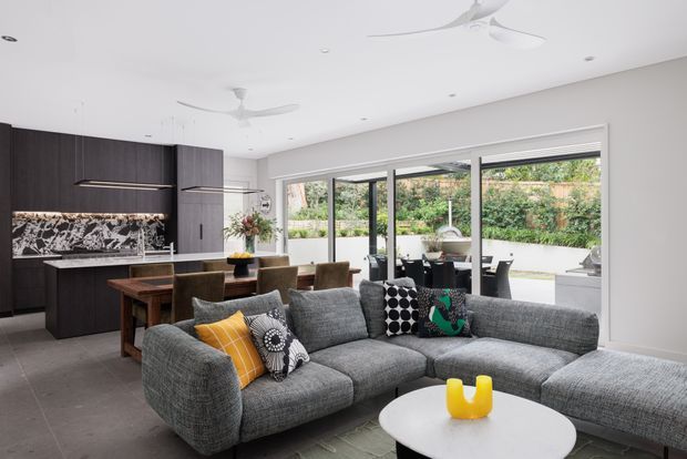 Open-plan contemporary living and dining area with a dark timber kitchen at the rear, a large grey sectional sofa in the foreground, and a dining table beside full-height glazed doors opening to a landscaped outdoor area. The room has a white ceiling with recessed lighting and ceiling fans, large grey floor tiles, and a striking black-and-white patterned kitchen splashback. Bright natural light enters through the extensive glazing.