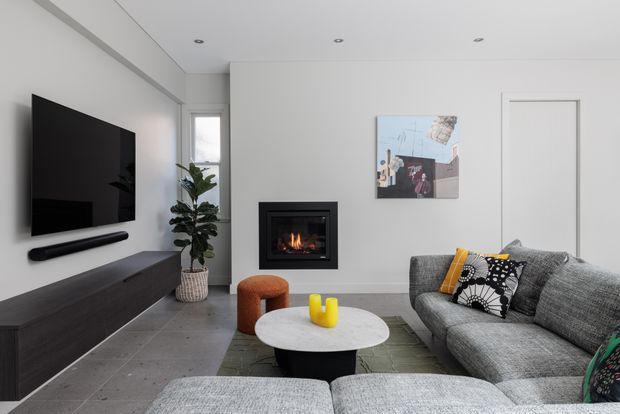 Contemporary living room with a large grey sectional sofa, wall-mounted television, low dark timber media cabinet, inset fireplace, round coffee table, and patterned cushions. The room has white walls and ceiling, grey concrete-look flooring, recessed downlights, and a narrow window with indoor planting. The atmosphere is clean, modern, and softly warm from the fireplace.