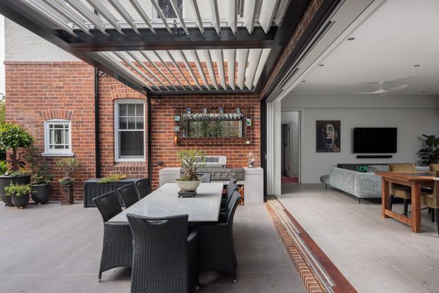 A covered outdoor dining terrace opens directly into a contemporary living room through wide glazed sliding doors. The terrace features a long dining table with dark woven chairs, brick walls, potted plants, and a striking overhead pergola with white adjustable louvres and dark structural framing. Inside, a pale sofa, television, timber dining furniture, artwork, and light flooring are visible. The space has a refined indoor–outdoor connection and a warm brick-and-neutral palette.