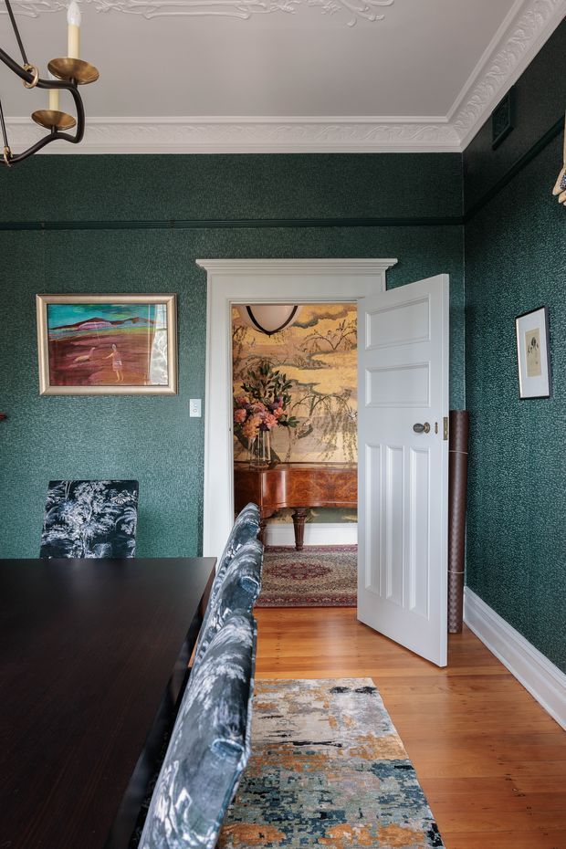 A richly decorated traditional dining room with deep green textured walls, ornate white ceiling cornices and decorative plasterwork, and warm timber flooring. A dark dining table with patterned upholstered chairs occupies the foreground. An open white paneled door reveals an adjoining room with a classical sideboard, floral artwork or wallpaper, and a patterned rug. Framed artwork and a brass-toned chandelier add refined vintage character.