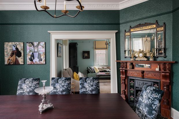 A richly decorated formal dining room with deep green textured walls, ornate white cornice and door trim, a dark timber dining table, patterned upholstered dining chairs, and a carved traditional timber fireplace mantel with a large decorative mirror. A living room is visible through the open doorway, and a classic suspended chandelier hangs overhead.