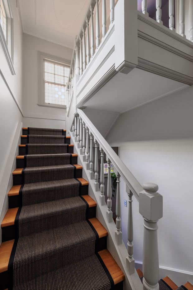A tall, narrow interior stairwell with a timber staircase, dark patterned carpet runner, and white painted timber balustrades. The staircase turns upward beside a landing, with traditional turned spindles, moulded trim, a decorative newel post, white walls, and a leaded or multi-pane window providing natural light.
