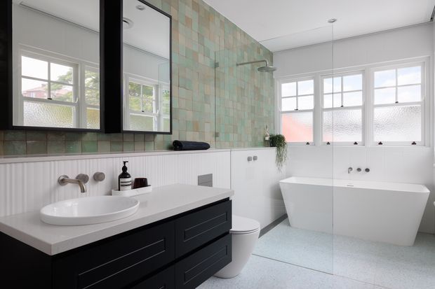Bright contemporary bathroom with a freestanding white bathtub, wall-mounted shower, floating dark vanity with an integrated white benchtop and basin, and large black-framed mirrors. Pale green and beige tiled walls contrast with white vertical wall panelling, frosted multi-pane windows, and a light terrazzo-like floor.