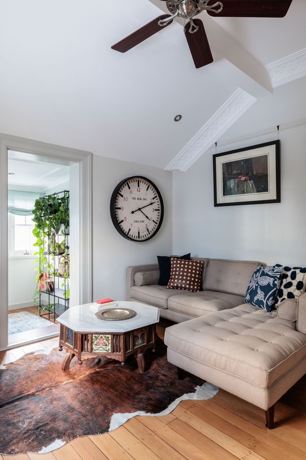 A bright, traditionally styled living room with a large beige sectional sofa, patterned accent cushions, an ornate octagonal coffee table with a pale stone top, and a brown hide rug over timber flooring. The room has pale grey walls, elaborate white decorative cornices, a dark timber ceiling fan, framed artwork, and a large round wall clock. An open doorway leads to a sunlit plant-filled adjoining room.