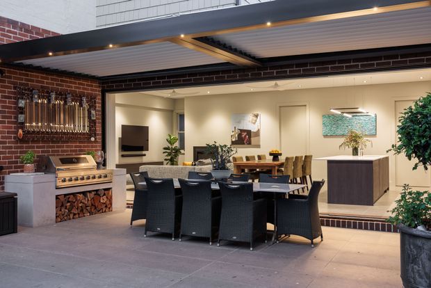 A covered outdoor entertaining area opening into a refined indoor dining and living space. The patio features a large dining table with black woven chairs, a built-in barbecue with timber log storage, exposed red-brick walls, a slatted soffit with recessed lighting, and tiled or concrete-look flooring. The adjoining interior includes a dining table, cabinetry, artwork, and warm neutral finishes, creating a sophisticated indoor-outdoor entertaining setting.
