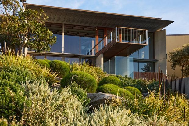 Exterior view of a contemporary multi-level house set into a landscaped sloping site. The building features a broad low-pitched roof overhang, extensive floor-to-ceiling glazing, warm timber soffits and structural elements, projecting balconies with slim metal balustrades, and a combination of dark and light rendered surfaces. Native-style planting and large rocks form the foreground landscape.