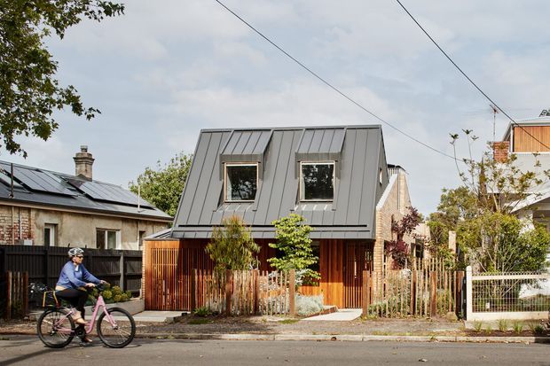 Street-facing view of a compact contemporary residential house in an established urban neighbourhood. The home has a steep, folded standing-seam metal roof and upper facade, warm vertical timber screening and cladding at ground level, dark-framed windows, a small planted front garden, and a simple pedestrian entry. Adjacent homes, overhead utility wires, and a cyclist are visible in the streetscape.