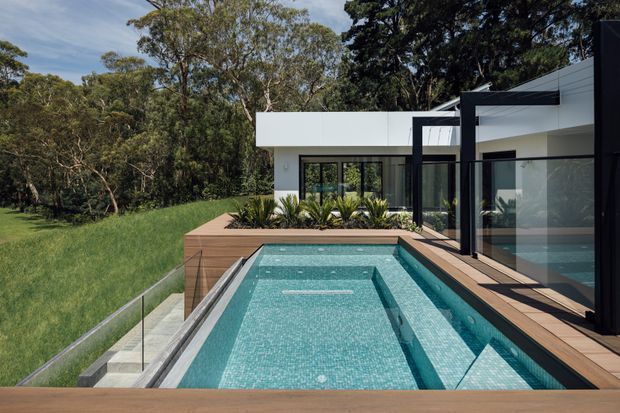 A contemporary flat-roofed house set within a leafy landscape, viewed from an elevated timber deck beside a rectangular swimming pool. The pool has pale blue mosaic tiling, integrated steps and a shallow ledge, with frameless glass balustrading along the lawn edge. White rendered walls, dark aluminium-framed glazing, bold black structural framing and timber decking define the architecture.