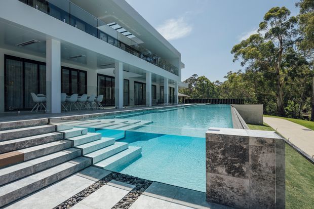 A contemporary multi-level residence with extensive floor-to-ceiling glazing, white structural columns, covered outdoor entertaining areas, and a large infinity-edge swimming pool. Broad stone steps descend into the pool, which is bordered by dark stone paving and a manicured lawn, with mature trees beyond the property.