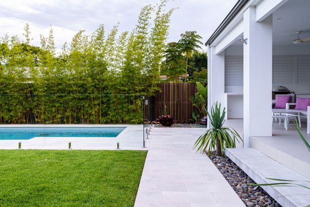 Bright contemporary outdoor living area beside a rectangular swimming pool, with a pale paved terrace, manicured lawn, bamboo screening, timber fencing, and a white pavilion-like structure with deep covered openings and outdoor seating. A landscaped gravel garden bed with architectural plants runs along the building.