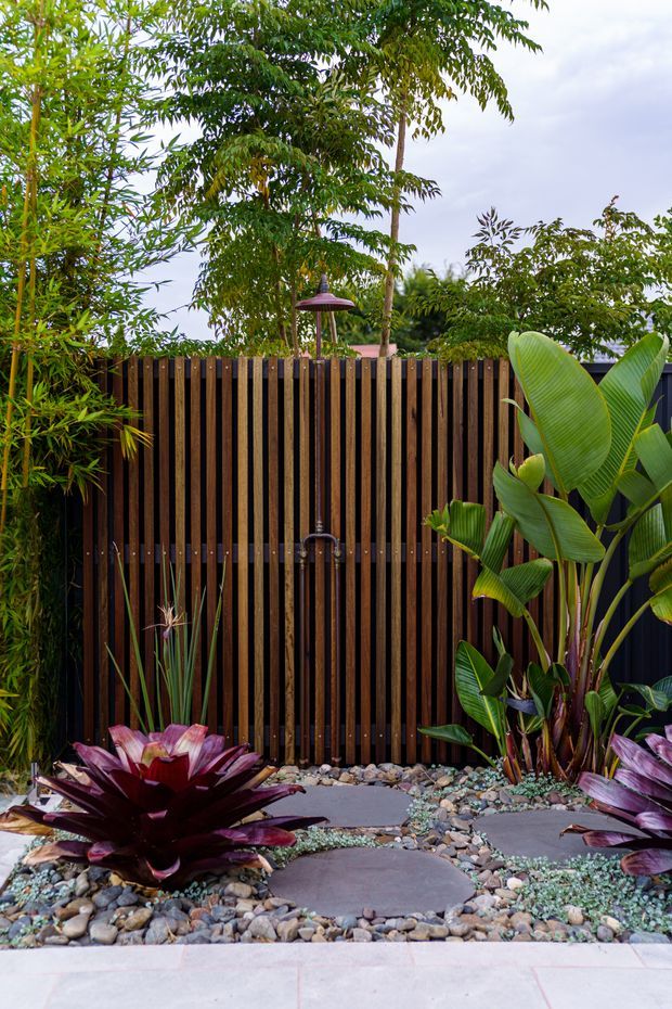 A lush landscaped courtyard or garden area with a vertical dark timber slat privacy fence, tropical foliage, bromeliads, bamboo, decorative river pebbles, and irregular stepping stones. A small wall-mounted outdoor shower or fixture is centered on the fence, with a slim purple outdoor light above it. The scene has a refined tropical resort-like character and a carefully designed low-maintenance garden.