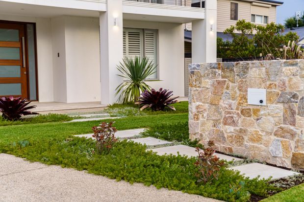 A contemporary residential facade viewed from the street, featuring a white rendered multi-level entry with broad columns, a timber-and-glass front door, louvred windows, and a landscaped front garden. A low wall of irregular beige and grey stone forms the foreground boundary, with concrete stepping slabs and sculptural planting leading to the entrance.