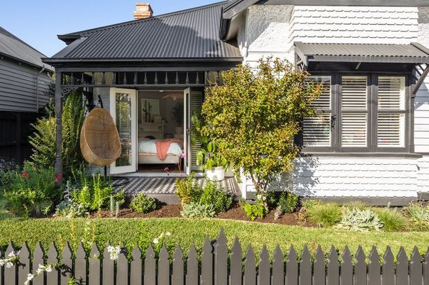 Front exterior of a compact cottage-style home with a dark corrugated metal roof, white weatherboard cladding, a covered entry porch, timber-framed glazing, shutters, and a landscaped garden behind a dark picket fence. A hanging chair is visible on the porch.