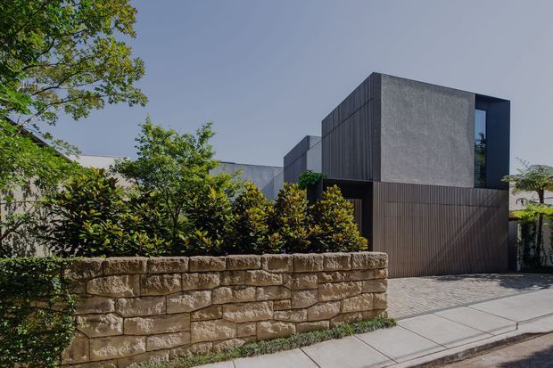 Street-facing view of a contemporary multi-level residence with strong rectilinear geometric forms, dark vertical slatted cladding, textured grey rendered walls, and a large recessed upper-level window. A substantial natural-stone retaining wall, landscaped screening plants, and a paved driveway and footpath define the frontage.