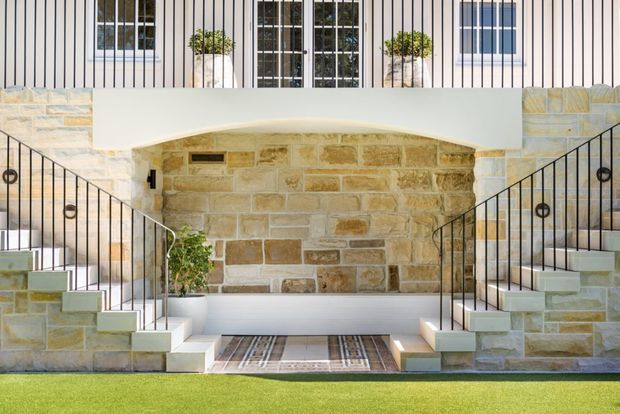 Symmetrical exterior entry facade with twin staircases leading to a recessed sandstone-clad lower-level entrance. Black metal balustrades with circular details frame the stairs, while a white rendered upper facade features narrow vertical cladding lines, gridded glazing, and potted greenery. A manicured lawn sits in the foreground.
