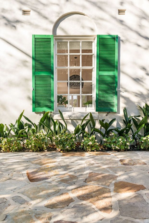 Exterior architectural detail of a light-coloured rendered wall with a tall multi-pane window, vivid green louvred shutters, and a decorative curved hood above the window. Low planting borders the wall, with irregular natural stone paving in the foreground. The window reveals a softly styled interior beyond.