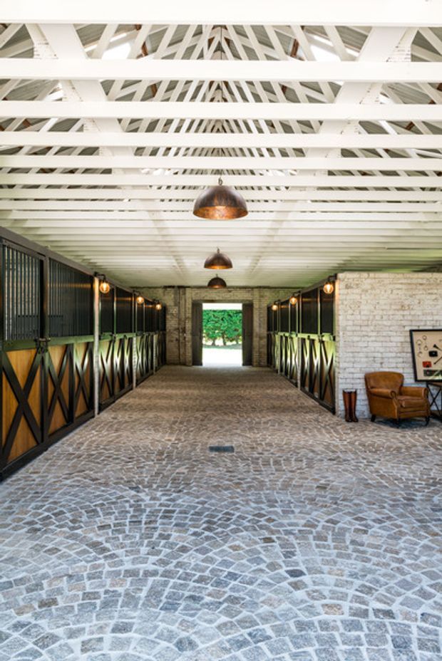 A symmetrical covered equestrian stable corridor with rows of dark metal-and-timber horse stalls on both sides. The space has a white painted exposed timber truss and beam roof structure, hanging pendant lights with bronze-toned shades, pale exposed brick walls, and a patterned cobblestone floor. A leather armchair and framed artwork sit near the right wall, while an open doorway at the far end leads outside.