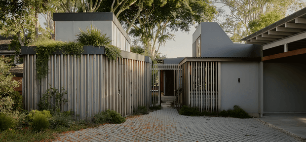 Exterior view of a contemporary residential property with several compact pavilion-like structures arranged around a paved entry courtyard. The facade combines pale grey rendered walls, vertical timber or metal screening, dark roof forms, planted edges, and a covered carport or garage on the right. Mature trees and dense landscaping surround the driveway and entry path.