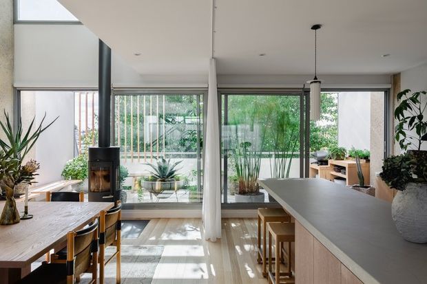 Bright open-plan residential interior combining a dining area and kitchen. The room features a long timber dining table with black-framed chairs, a central island with a dark grey benchtop and light timber cabinetry, extensive floor-to-ceiling glazing, indoor plants, a freestanding black wood-burning stove, and a light timber floor. A planted courtyard or garden is visible through the glazing.