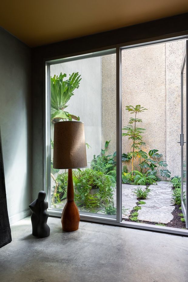 A moody, minimalist interior corner with a polished concrete floor, dark plastered walls, and a warm-toned ceiling. Full-height glazed sliding doors open onto a lush enclosed courtyard garden with stone stepping paths. A sculptural timber floor lamp and dark abstract figurine provide simple decorative accents.