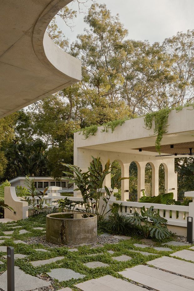 A landscaped courtyard or garden terrace framed by sculptural pale concrete architecture. The space features curved roof forms, a covered arcade with repeated arched openings, lush tropical planting, gravel garden beds, a circular concrete planter or water feature, and irregular light paving set into groundcover.