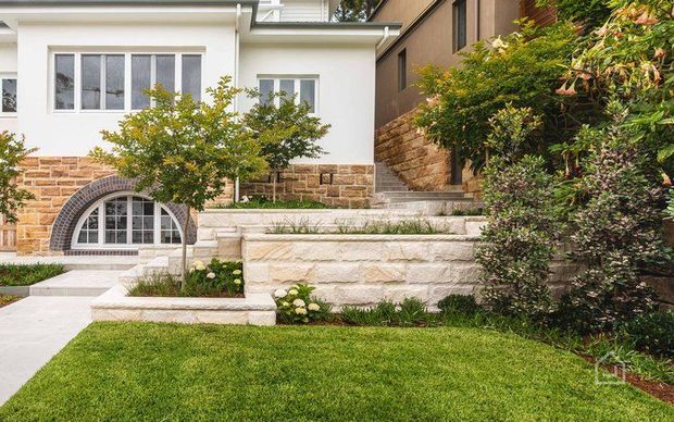 Exterior view of a landscaped residential property with a white rendered facade, arched lower-level window, stone masonry detailing, stepped retaining walls, paved walkway, lawn, and established shrubs and trees.