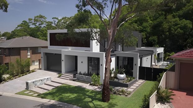 Exterior view of a contemporary multi-level residence with crisp white rendered walls, dark framed glazing, a flat roof, sheltered entry, integrated double garage, and landscaped front yard. The home sits in a suburban streetscape surrounded by mature trees and neighbouring houses.