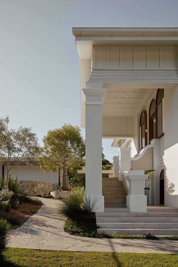 Exterior view of a refined white rendered house entrance, featuring a covered front porch with substantial square columns, decorative layered cornice detailing, a coffered ceiling, arched timber-framed windows, and broad stone steps. A landscaped path with native-style planting leads to the entry, with a low stone boundary wall visible beside the property.