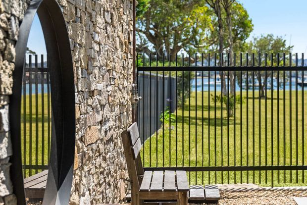 Exterior view of a stone-clad entry or boundary structure beside a black metal picket fence and gate. A small timber bench sits on a gravel-paved area, with a manicured lawn and blue water visible beyond the fence. The image has a bright, relaxed coastal or waterfront character.