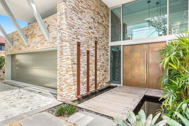 Contemporary residential entry facade featuring textured natural stone cladding, a broad integrated garage, tall glazed entry doors, exposed white roof beams, and a timber footbridge over a shallow water feature. Tropical planting and patterned paving soften the modern exterior.