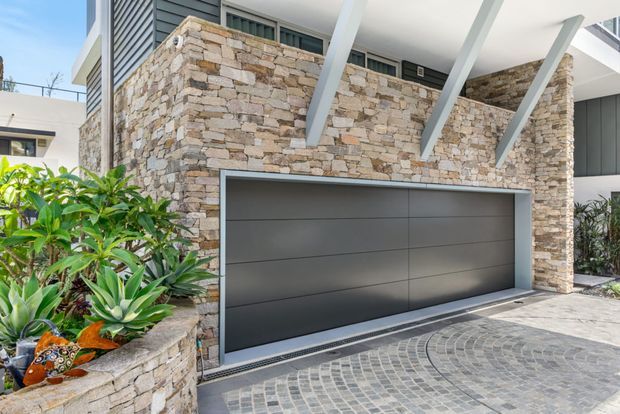 Contemporary residential facade featuring a wide dark grey sectional garage door set into a wall clad in irregular stacked natural stone. White structural roof beams project over the garage, with dark louvred detailing and upper glazing above. A curved cobblestone driveway, stone planter wall, and tropical landscaping complete the exterior composition.