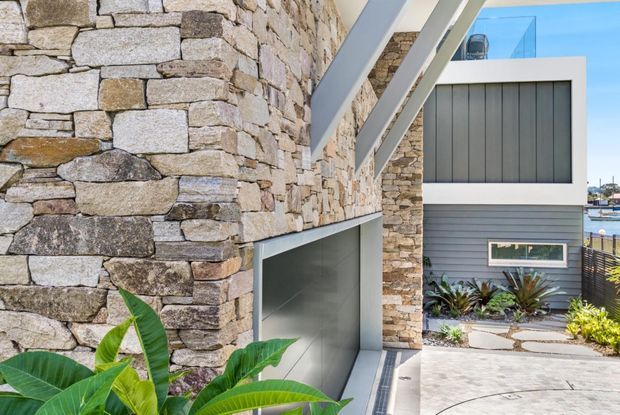 Exterior architectural view of a contemporary coastal residence, focusing on a robust stacked-stone wall, large integrated garage door, white structural framing, and grey horizontal cladding. A paved driveway and planted entry garden lead toward the house, with glimpses of water and neighboring buildings in the background.