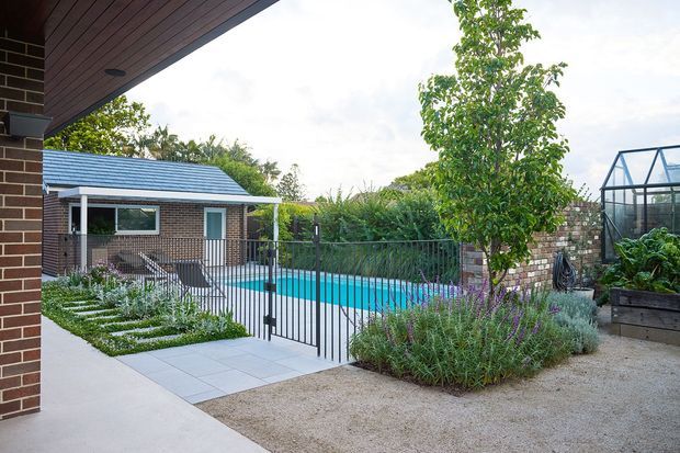 A landscaped residential courtyard viewed from beneath a covered outdoor area, featuring a turquoise swimming pool enclosed by a black metal fence, a small brick pool house with a blue metal gabled roof, paved paths, gravel garden areas, flowering plants, and a glass greenhouse.