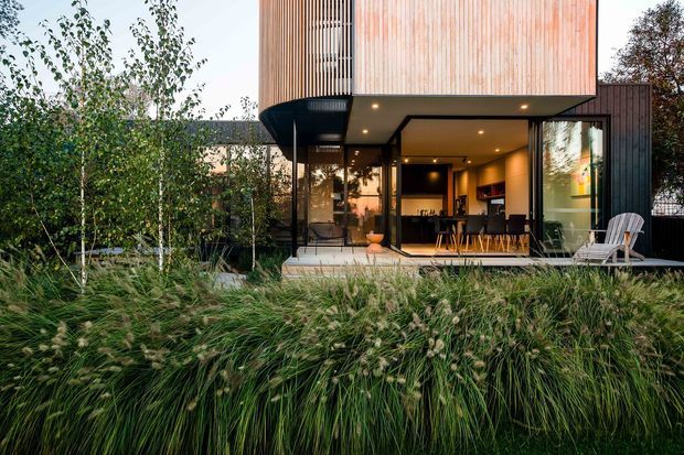Exterior view of a contemporary two-storey home set within landscaped planting. The upper level features warm vertical timber cladding and a projecting box-like form, while the lower level opens to a covered outdoor living area with extensive glazing, a kitchen or dining space, and dark cladding. Ornamental grasses and slender trees form the foreground landscape.