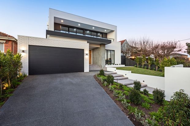 Contemporary two-storey suburban residence viewed from the street, featuring a white rendered and masonry facade, a large dark garage door, upper-level balcony with recessed downlights, broad glazing, and a landscaped front garden with stepped entry path and lawn.