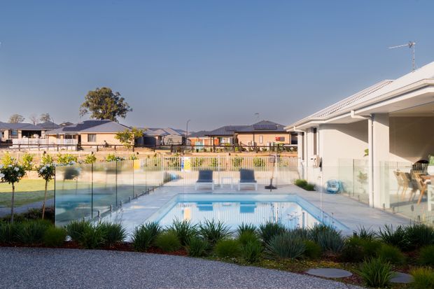 A landscaped residential pool area viewed from an outdoor patio or terrace, with a rectangular swimming pool enclosed by glass fencing, low ornamental planting, and a modern single-storey house with a covered outdoor living area on the right. Neighbouring suburban homes and a clear blue sky are visible beyond the property.