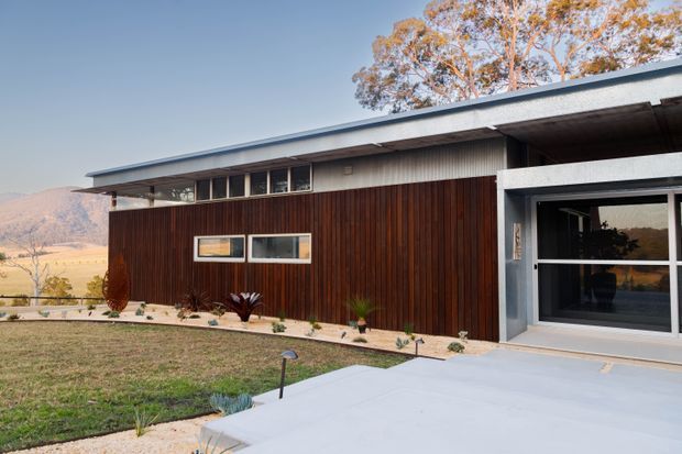 Single-storey contemporary residence viewed from the front, with a long low-slope roof, dark vertical timber-clad facade, exposed concrete entry surround, clerestory-style glazing, and a broad pale concrete pathway and terrace. The home sits in a dry rural or semi-rural landscape with hills and mature trees in the background.