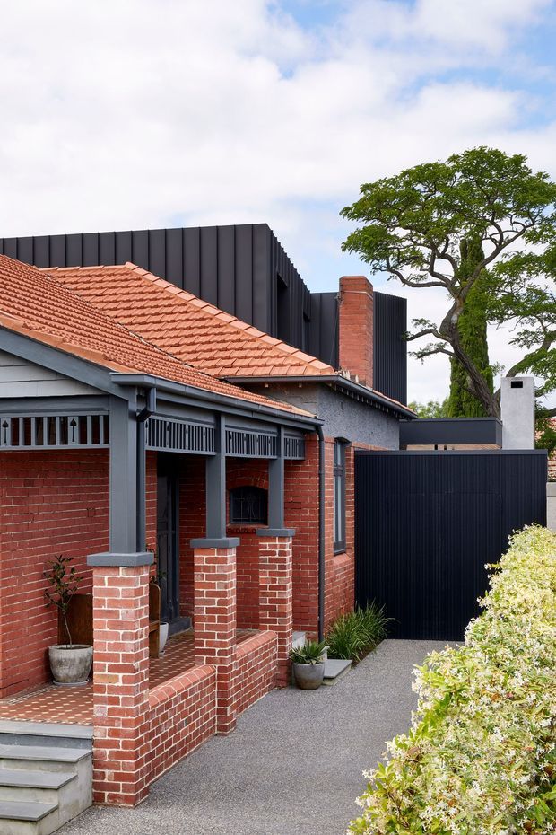 Street-facing view of a renovated brick house combining a traditional red terracotta-tiled roof and covered front veranda with a contrasting contemporary dark vertical-metal extension, brick chimney, dark gate, paved entry path, and established landscaping.