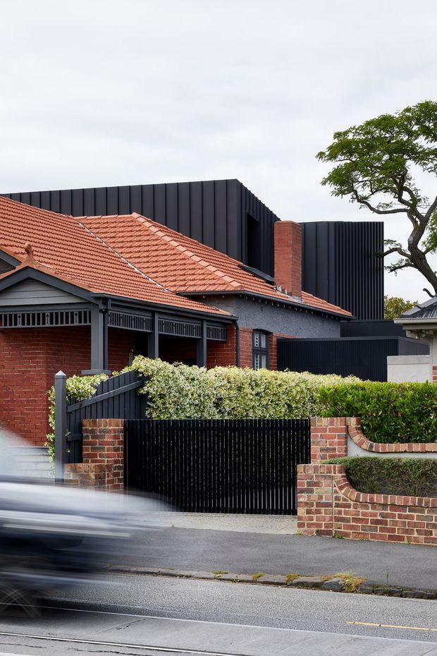 Street-level view of a renovated suburban brick residence combining a traditional red terracotta tiled roof and brick facade with a bold contemporary black vertical-clad upper extension. The property features dark metal fencing and gates, a covered front verandah with decorative screening, clipped hedges, stepped brick garden walls, and a mature street tree. A blurred passing vehicle appears in the foreground.