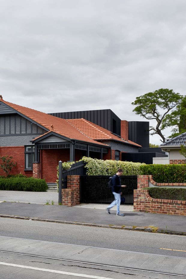 Street-facing view of a residential dwelling combining a traditional brick and terracotta-tiled gabled house with a contemporary dark vertically clad extension. The property has a black metal front gate, brick boundary walls, hedges, and mature landscaping, photographed from across a street with tram tracks visible in the foreground.