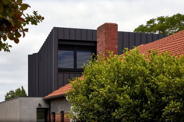 Exterior view of a contemporary dark-clad upper-storey addition rising behind a traditional red terracotta-tiled roof. The addition features vertically ribbed dark metal cladding, a large horizontal louvred window, and a prominent exposed red-brick chimney, with mature hedging partially obscuring the lower facade.