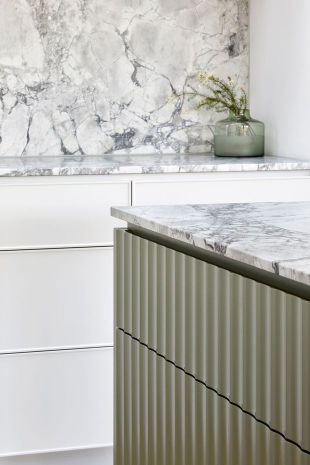 Close-up view of a refined kitchen featuring a veined white-and-grey marble splashback and countertop, white framed cabinetry, and a sculptural fluted island in a muted sage-grey finish. A translucent green decorative vessel with foliage sits on the rear benchtop.