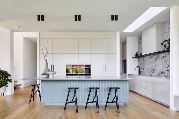 Bright contemporary kitchen with a large white island featuring a vertically fluted front, three black stools, extensive white cabinetry, integrated appliances, and a marble-look backsplash with open shelving. Light timber flooring, recessed lighting, indoor plants, and a large skylight create an airy, refined interior.