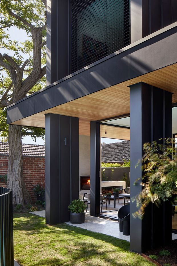A close exterior view of a contemporary residential addition with dark charcoal cladding, strong rectilinear forms, deep overhangs, and warm timber soffits. Full-height glazing opens toward a small landscaped courtyard, with a visible outdoor fireplace and seating area. A mature tree, lawn, brick boundary wall, and neighbouring tiled roof frame the setting.