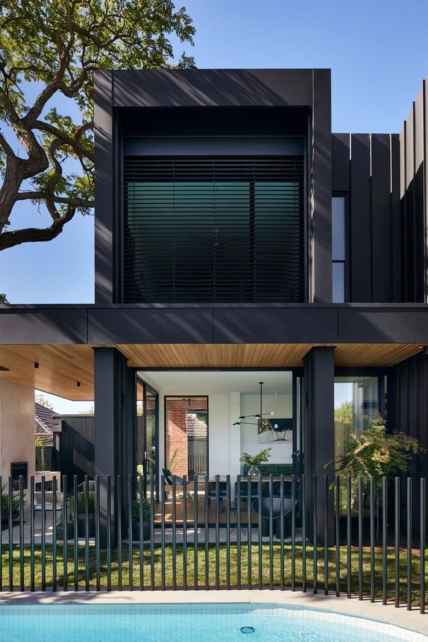 A contemporary multi-level residential facade viewed from the pool area. The dark charcoal cladding and strong rectilinear forms frame a recessed upper window with horizontal louvres, while the lower level opens to a covered dining or living area with extensive glazing. Warm timber soffits contrast with the dark exterior, and a vertical metal fence separates the house from the bright swimming pool in the foreground.